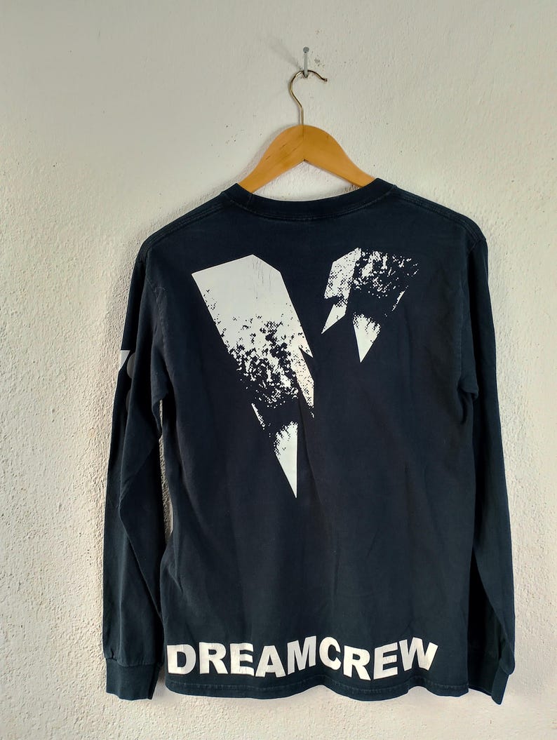 Pode incluir: Camiseta preta de manga comprida com um design gr&aacute;fico branco nas costas. O design inclui um raio e o texto "DREAMCREW" em letras brancas. A camiseta est&aacute; pendurada em um cabide de madeira.