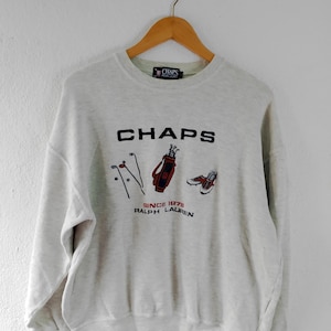 Puede incluir: Una sudadera gris claro con un logotipo "CHAPS" negro y un gráfico con temática de golf. El gráfico incluye una bolsa de golf, palos de golf y zapatos de golf. El texto "SINCE 1978 RALPH LAUREN" está impreso debajo del gráfico.