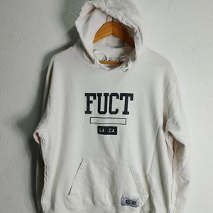 May include: Cream-colored hoodie with a hood and a front pocket. The word "FUCT" is printed in bold black letters across the chest, with "LA CA" below. A small tag with text is on the lower right.