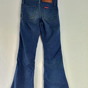 May include: Blue denim bell-bottom jeans with a faded appearance. The jeans have two back pockets and a small red tag. A brown leather patch is visible at the waistband. The jeans are hanging on a black hanger.