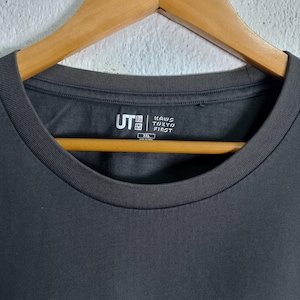Puede incluir: Camiseta gris oscuro con cuello redondo. La camiseta presenta el texto "KAWS TOKYO FIRST" y el logotipo "UT". La prenda est&aacute; colgada de una percha de madera.