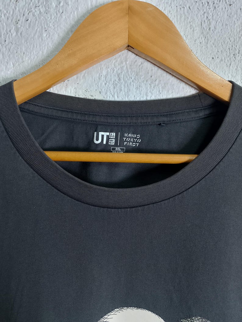 Puede incluir: Camiseta gris oscuro con cuello redondo. La camiseta presenta el texto "KAWS TOKYO FIRST" y el logotipo "UT". La prenda est&aacute; colgada de una percha de madera.