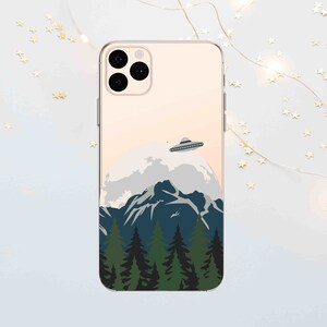 Op de afbeelding: Een telefoonhoesje met een afbeelding van een UFO die over een bergketen vliegt met een bos op de voorgrond. Het hoesje is transparant en heeft een gouden rand.