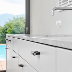 May include: White kitchen cabinets with black drawer pulls. The cabinets are made of wood and have a smooth finish. The drawers are all closed and the pulls are centered on the drawers. The cabinets are in a modern kitchen with a marble countertop and a stainless steel sink.