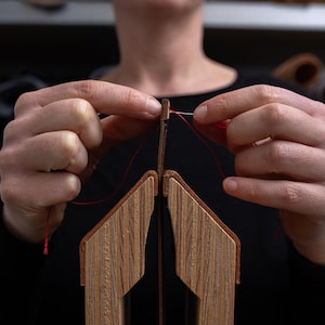 Può includere: Primo piano di uno strumento artigianale in legno con un filo rosso che lo attraversa. Lo strumento ha un design geometrico. La persona che tiene lo strumento indossa una maglietta nera. Lo sfondo è sfocato.