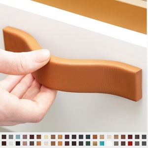 May include: A curved, tan leather cabinet pull with a textured surface. The pull is mounted on a white cabinet door.