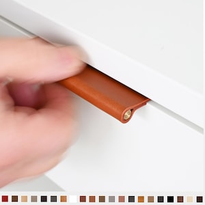 May include: A close-up of a brown leather drawer pull with a brass accent. The pull is mounted on a white cabinet door.