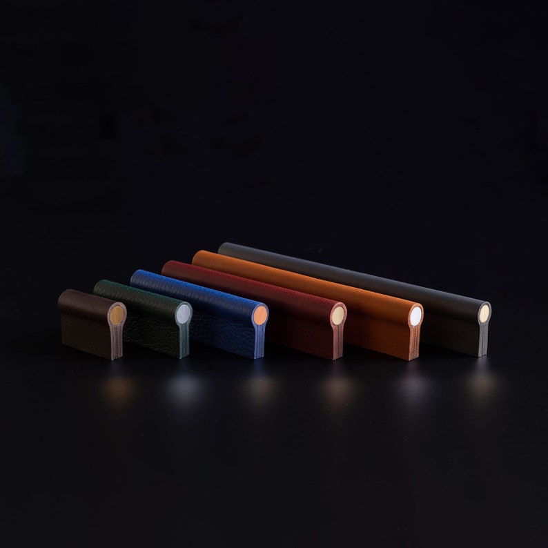 May include: A set of eight leather drawer pulls in various colors, including black, dark green, light blue, dark blue, burgundy, brown, tan, and gray. Each pull has a rounded end and a metal accent.