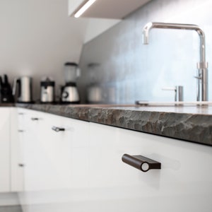 May include: White kitchen cabinets with brown cabinet pulls. The countertop is made of a dark, textured material. A silver faucet is visible in the background.