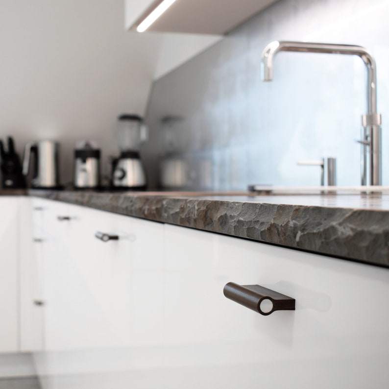May include: White kitchen cabinets with brown cabinet pulls. The countertop is made of a dark, textured material. A silver faucet is visible in the background.