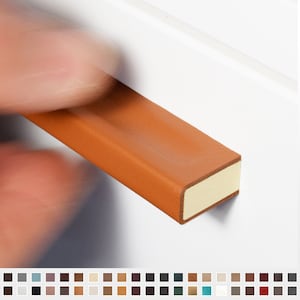 Könnte beinhalten: Nahaufnahme eines hellbraunen Leder-Schubladengriffs mit cremefarbenem Rand. Der rechteckige Griff wird von einer verschwommenen Hand gehalten. Der Text "RIVA-MINI" ist im Hintergrund zu sehen.