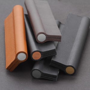 May include: Close-up of five leather cabinet pulls in different colors. The pulls are made of leather wrapped around a metal core. The colors are brown, black, and gray.