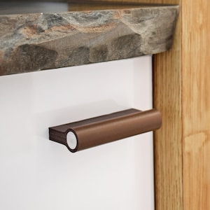 May include: A brown leather cabinet pull with a silver metal accent. The pull is mounted on a white cabinet door.