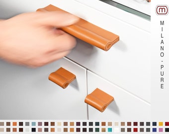 May include: Three leather cabinet pulls in various sizes. The pulls are a light brown color and are folded over at the ends. The pulls are mounted on a white cabinet door. The text "MILANO - PURE" is visible in the upper right corner of the image.