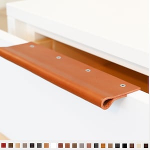 Può includere: Primo piano di una maniglia per cassetto in pelle marrone con viti argentate. La maniglia è montata su un frontale del cassetto bianco.