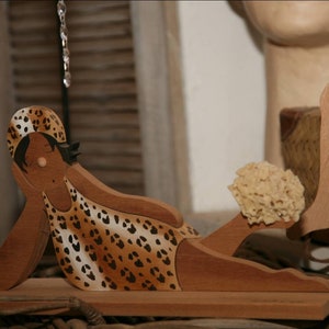 May include: Wooden figurine of a person wearing a leopard print swimsuit and a hat, lying on a wooden base. The figurine is holding a sponge in one hand.