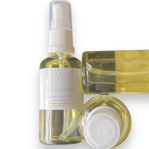 May include: Three bottles of clear cleansing oil with white pump tops. The bottle in the centre has a white label with the text "MONEOPURA Cleansing Oil Oil to Milk www.moneopura.com".