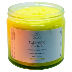 Può includere: Un barattolo di vetro di scrub corpo alla curcuma giallo con un'etichetta bianca su cui è scritto "Turmeric Scrub" e "Smoothing Body Scrub", con gli ingredienti elencati come "Turmeric, Earthy & Lemon". L'etichetta include anche il nome del marchio "Monegaura" e l'indirizzo del sito web "www.monegpura.com".