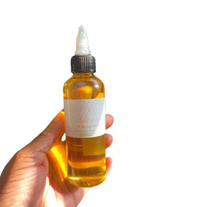 May include: A clear glass bottle filled with amber-colored liquid, held in a hand. The bottle has a white label with the text "MONEOPURA Fenugreek Extreme Growth Oil". The bottle has a black cap and a clear applicator tip. The background is white.