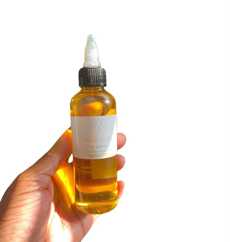May include: A clear glass bottle filled with amber-colored liquid, held in a hand. The bottle has a white label with the text "MONEOPURA Fenugreek Extreme Growth Oil". The bottle has a black cap and a clear applicator tip. The background is white.