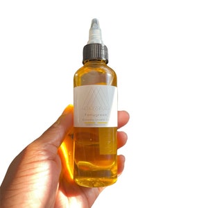 May include: A clear glass bottle filled with amber-colored liquid, held in a hand. The bottle has a black cap and a white label with the text "Moneopura Fenugreek Extreme Growth Oil". The background is white.