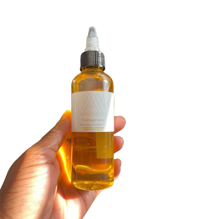 May include: A clear glass bottle filled with amber-colored liquid, held in a hand. The bottle has a black cap and a white label with the text "Moneopura Fenugreek Extreme Growth Oil". The background is white.