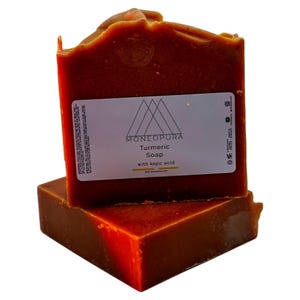 Può includere: Due saponette di curcuma di colore arancio-marrone con l'etichetta "Moneopura Turmeric Soap with Kojic Acid" sulla saponetta superiore.