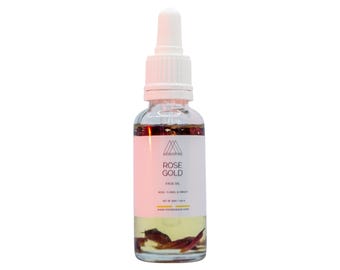 May include: A clear glass bottle with a white dropper top filled with a light yellow liquid and rose petals. The label reads "ROSE GOLD FACE OIL".