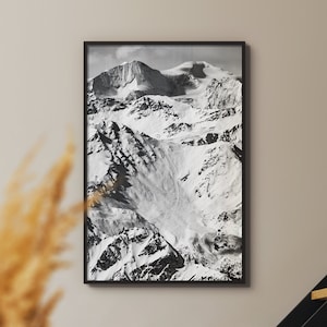 Op de afbeelding: Zwart-witfoto van een besneeuwde bergketen. De foto is ingelijst in een zwart frame en hangt aan een lichtbeige muur.
