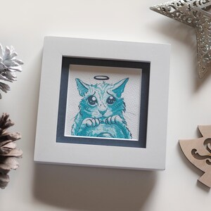 May include: A framed illustration of a blue cat with a halo, looking sad with big eyes. The cat is drawn in a cartoon style and has a white background.