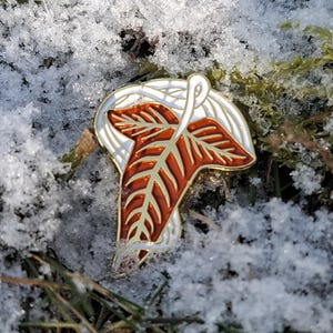 May include: A silver-colored pin with a detailed, stylized leaf design in shades of brown and orange. The pin is set against a backdrop of snow-covered ground with patches of green grass and moss.