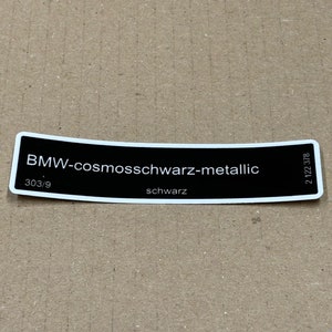 Puede incluir: Una pegatina en blanco y negro con el texto "BMW-cosmoschwarz-metallic" y los números "303/9" y "2 122 578". La pegatina también tiene la palabra "schwarz" impresa.