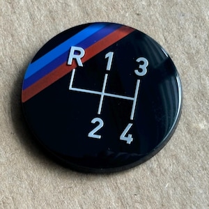 May include: A black, round gear shift knob with white numbers and a gear pattern. The top left corner features a design with red, blue, and orange stripes. The knob is likely for a car.