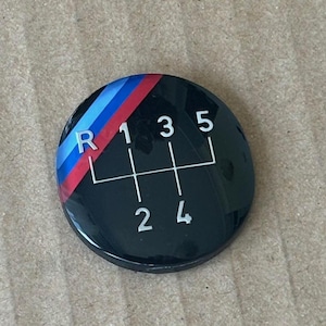 Könnte beinhalten: Ein runder schwarzer Aufkleber mit einem weißen Schaltmuster und den Buchstaben R, 1, 2, 3, 4 und 5. Das Schaltmuster hat oben einen blau-rot-weißen Streifen.