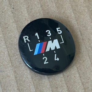 Peut inclure: Un pommeau de levier de vitesses rond noir avec des lettres blanches et un logo BMW bleu et rouge. Les lettres indiquent "R 1 3 5 L M 2 4".