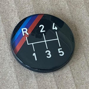 May include: A black round gear shift knob with a white gear shift pattern and a blue, red, and white stripe design.