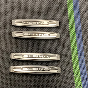 May include: Four silver metal car emblems with the word "ALPINA" in black lettering.