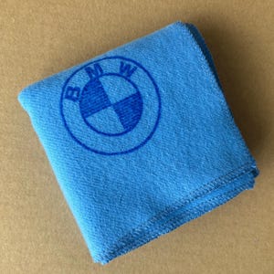 May include: A folded, light blue microfiber cloth featuring the BMW logo in the center. The logo is a blue circle with the BMW emblem in the middle. The cloth is made of a soft material and is likely used for cleaning.