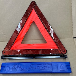 May include: A red reflective warning triangle with a black frame and a blue carrying case. The case has the text "Euro Werndreieck nach ECE R21 Warning Triangle Triangle de présignalisation Triengoll" and "DELTA 1" printed on it.