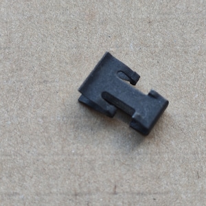 May include: A black plastic clip with a T-shaped design. The clip has two prongs on the top and a flat base.