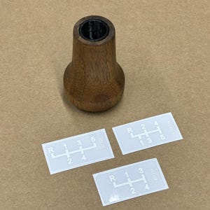 Puede incluir: Tres pegatinas blancas con n&uacute;meros y l&iacute;neas negras, que muestran diferentes patrones de cambio de marchas. Las pegatinas est&aacute;n sobre una superficie marr&oacute;n junto a una palanca de cambios de madera.