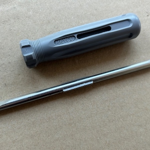 May include: A gray screwdriver with a black handle and the word "HEYCO" printed on it. The screwdriver has a flat head and a long, thin shaft.