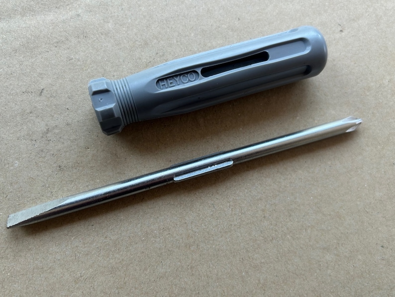 May include: A gray screwdriver with a black handle and the word "HEYCO" printed on it. The screwdriver has a flat head and a long, thin shaft.