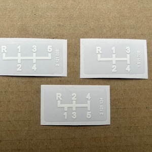 May include: Three white gear shift pattern stickers with black numbers and letters. The stickers have the numbers 1, 2, 3, 4, and 5, and the letter R. The stickers also have part numbers printed on them: 2 121 028, 2 121 134, and 1811 705.