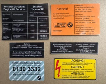 May include: A collection of six rectangular automotive warning and information stickers for BMW vehicles. The set includes black, orange, yellow, and silver labels featuring technical text in English, German, French, and Italian regarding oil, battery, cooling, and ignition systems.
