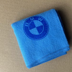 May include: A folded blue cloth with the BMW logo in the center. The logo is a blue circle with the letters "BMW" and a cross design in the middle. The cloth is folded neatly and has a textured appearance.