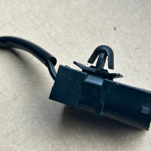 Może przedstawiać: Zbliżenie czarnego złącza elektrycznego z dołączonym czarnym przewodem. Złącze posiada klips i prostokątny korpus. Przewód jest pokryty czarną izolacją. Tło to jasnobrązowa powierzchnia.