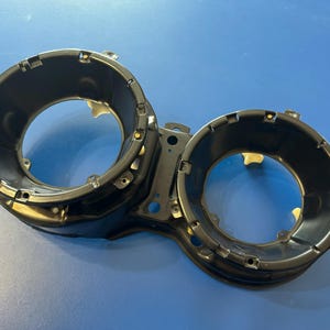 May include: Black automotive headlight housing with two circular openings. The housing is made of metal and has several mounting points. The background is a solid blue color.