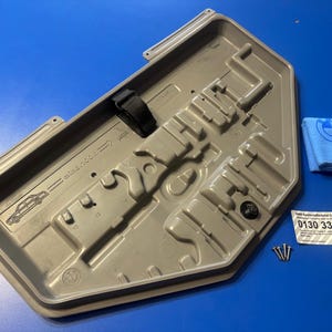 May include: A gray plastic car part with a black strap, a blue microfiber cloth with a logo, screws, and a label with a phone number. The car part has a complex molded design and a car silhouette.