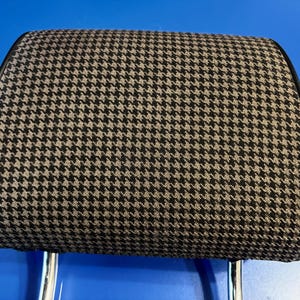 May include: A car headrest with a houndstooth pattern in black and tan. The headrest has a black leather trim and chrome support posts. The headrest is rectangular with rounded corners.